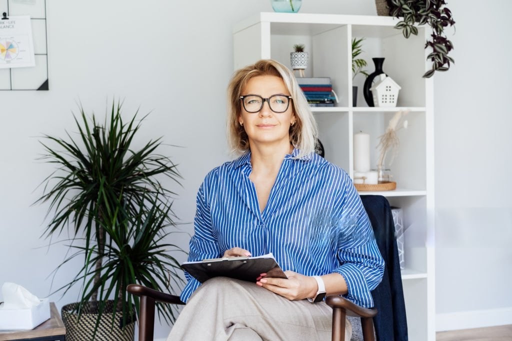 A confident woman wearing glasses and a blue striped shirt sits in a modern office space, holding a digital tablet. She appears calm and professional, with a bookshelf and potted plants in the background, suggesting a work or therapy environment.