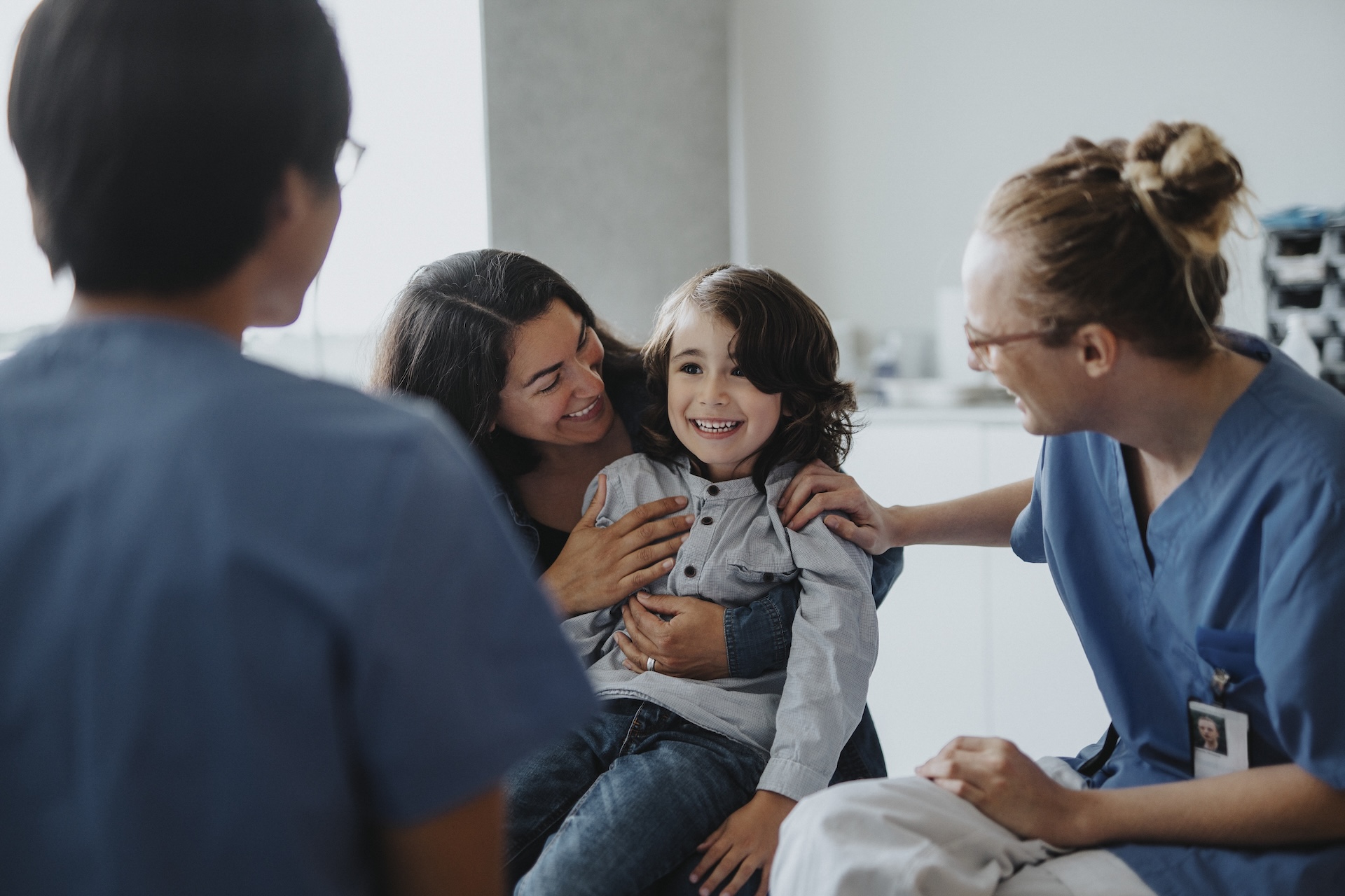 A happy child sits on an adult's lap, surrounded by smiling medical professionals. The group shares a lighthearted moment in a bright and caring clinical environment.