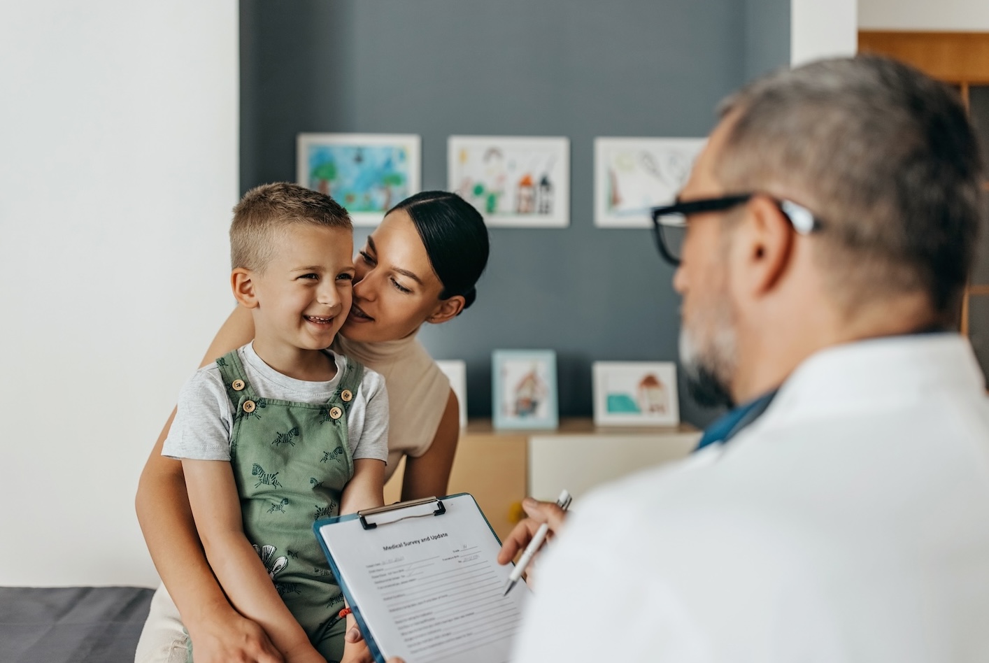 A young boy sits on his mother's lap, smiling happily while a doctor with a clipboard speaks to them. The room is decorated with children's artwork, creating a warm and welcoming atmosphere.