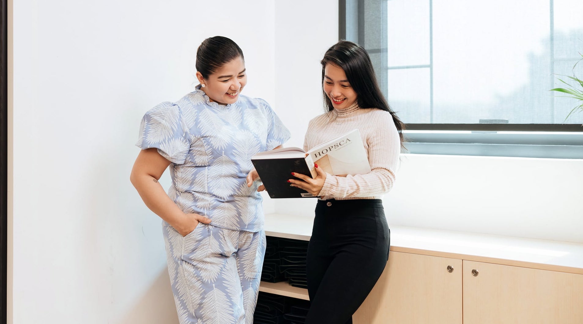 Two women stand together in a brightly lit room, smiling as they look at an open book titled 'HOPSCA.' One is wearing a light blue patterned outfit with puffed sleeves, and the other is dressed in a beige ribbed sweater and black pants.