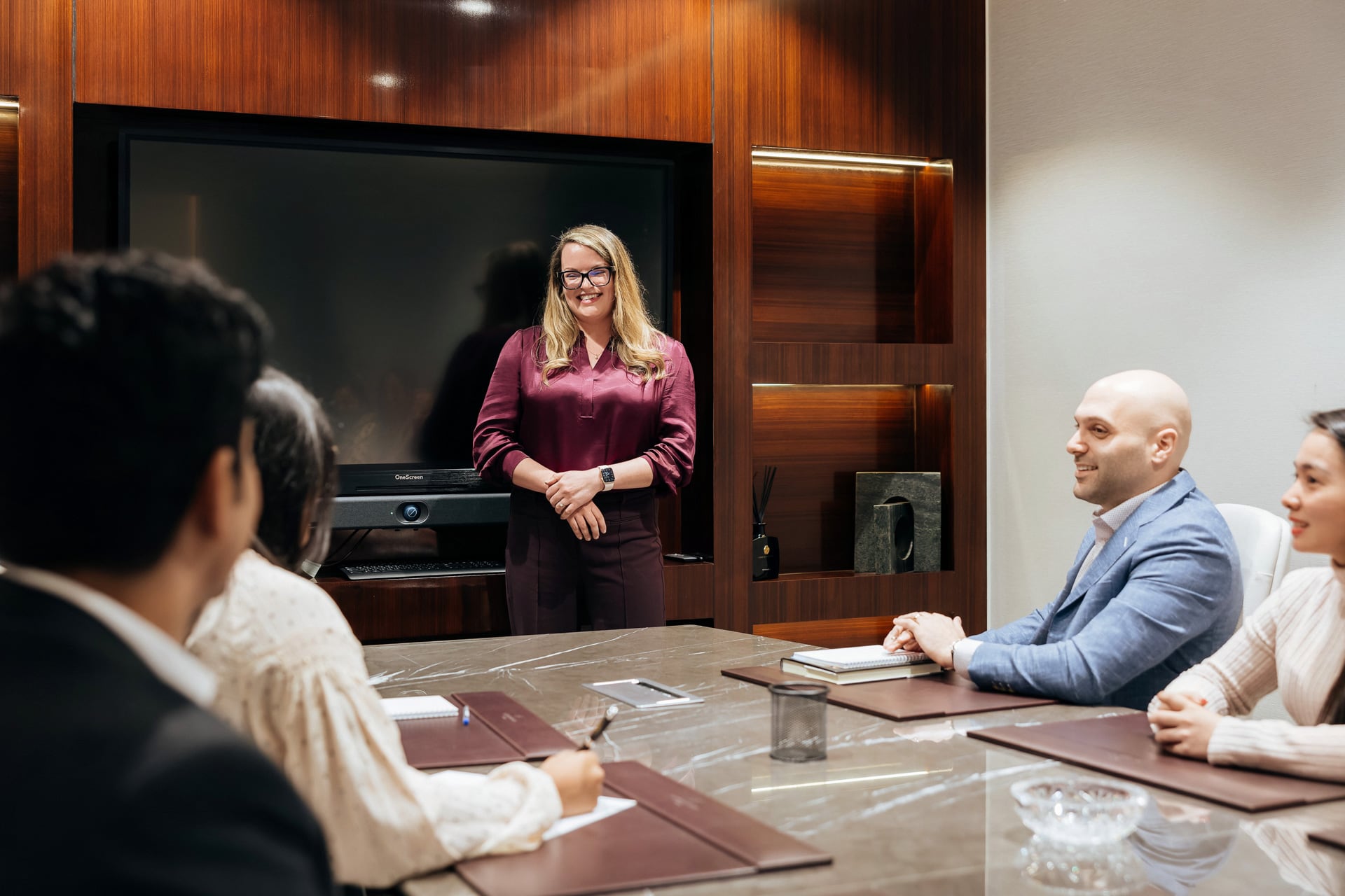 A woman stands at the front of a conference room near a large screen, smiling as she addresses a small group of seated professionals. The setting is modern, with polished wood paneling and a collaborative atmosphere.