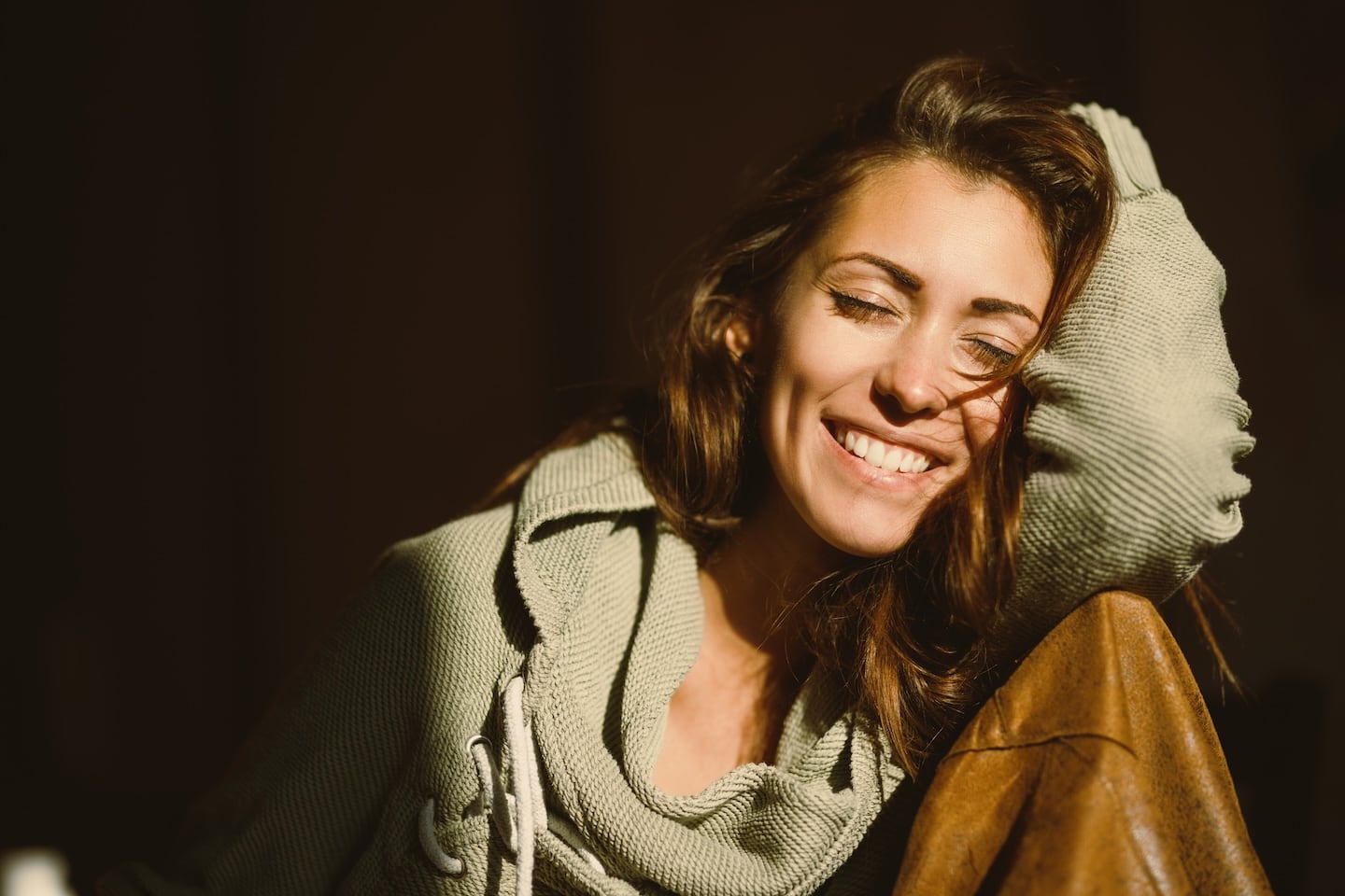 A cheerful woman with long brown hair smiles with her eyes closed, resting her head on her sweater-covered arm. Soft sunlight highlights her face, creating a cozy and serene mood against a dark backdrop.