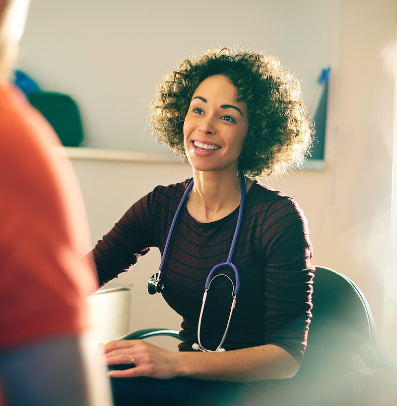 Smiling woman with curly hair and a stethoscope around her neck, seated and engaging warmly in conversation. She wears a maroon top, and the well-lit setting suggests a casual, supportive healthcare or clinical environment.