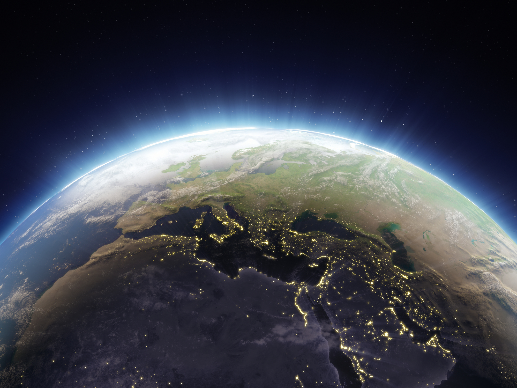 A view of Earth from space showing a sunrise over the Northern Hemisphere. City lights are visible across parts of Europe, the Middle East, and North Africa, contrasting with the dark night side of the planet. A soft blue glow outlines the horizon, with stars scattered in the background.
