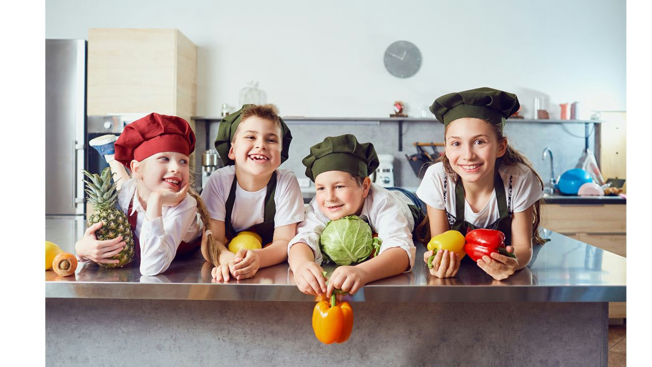 Kinder mit Kochmützen mit Gemüse in den Händen lehnen sich über eine Schneidefläche in einer Küche.