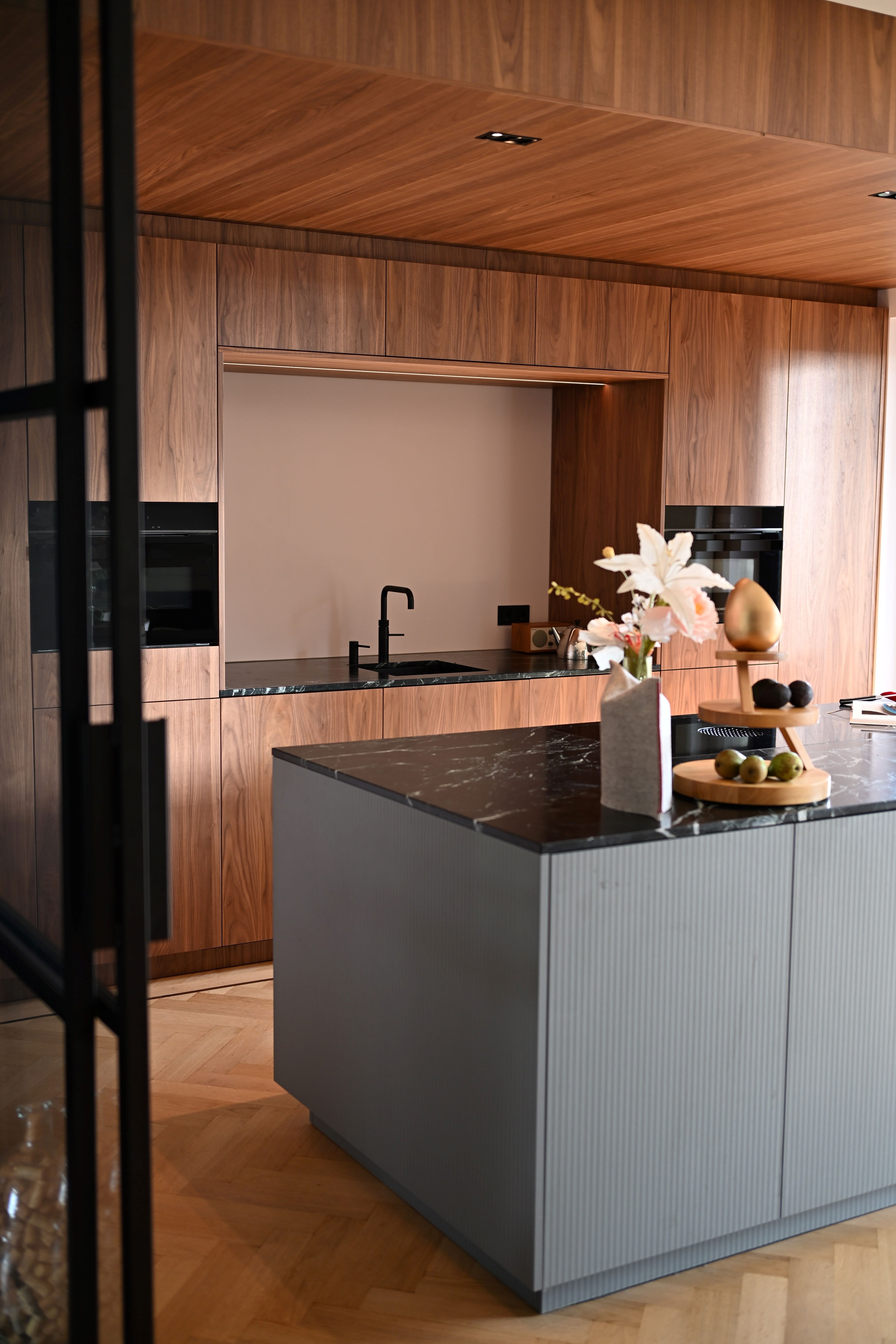 Moderne keuken met houten kasten, zwart marmeren werkblad en een vaas met bloemen en fruit op het eiland.