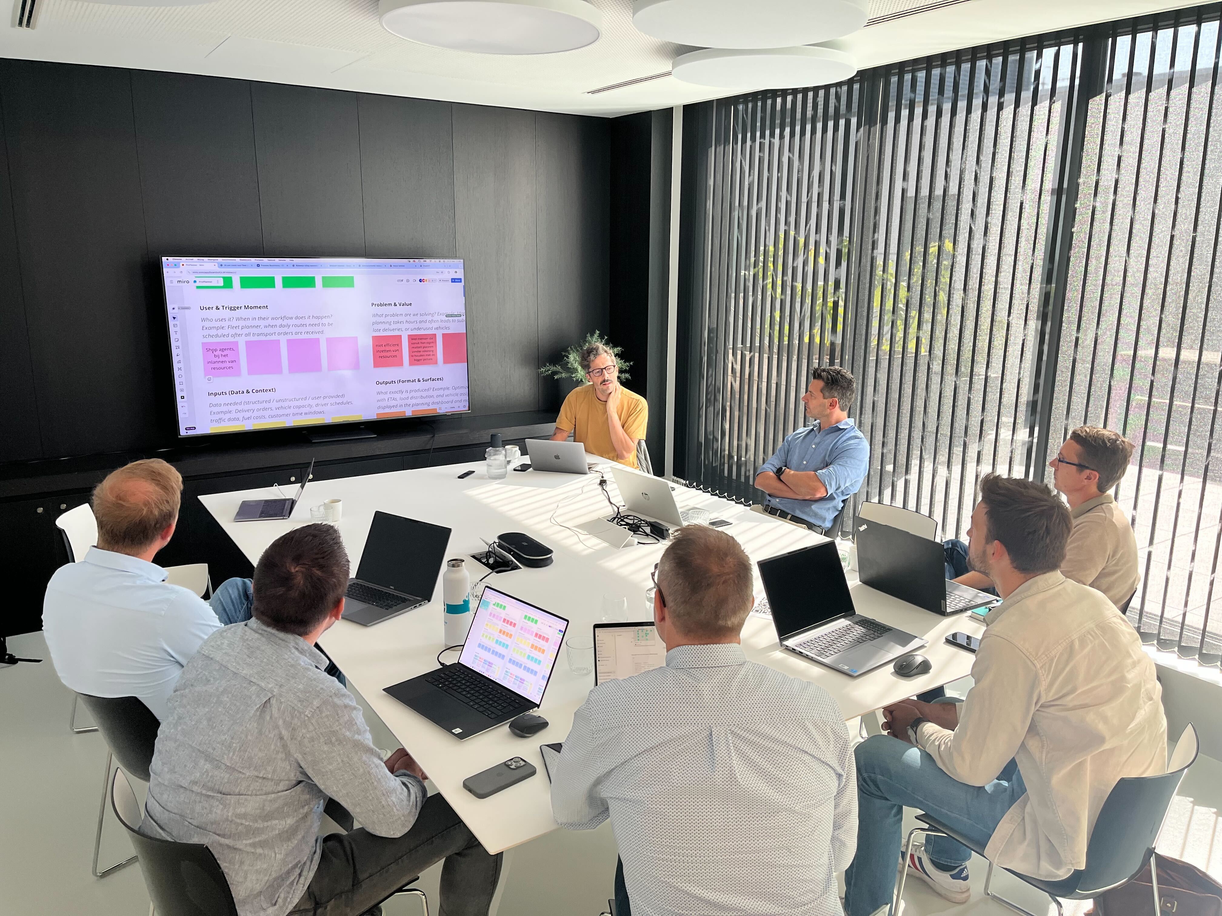 A group of professionals sits around a large white table in a modern office, focused on a presentation displayed on a large screen. The screen shows a project planning board with colored sticky notes, while laptops and water bottles are scattered across the table.