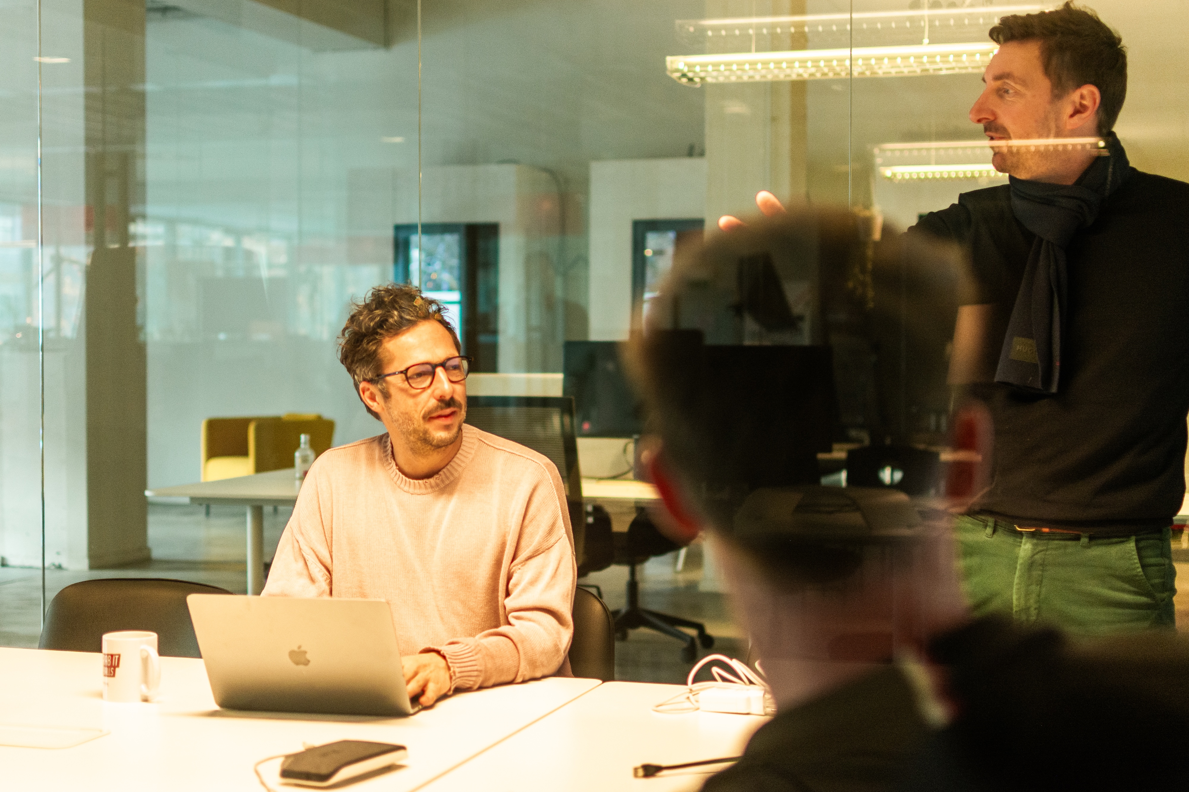 A man in glasses and a pale pink jumper sits with an open laptop at a white table in a warmly lit modern office, looking up at a standing colleague who gestures while talking, with glass partitions and desks behind them.