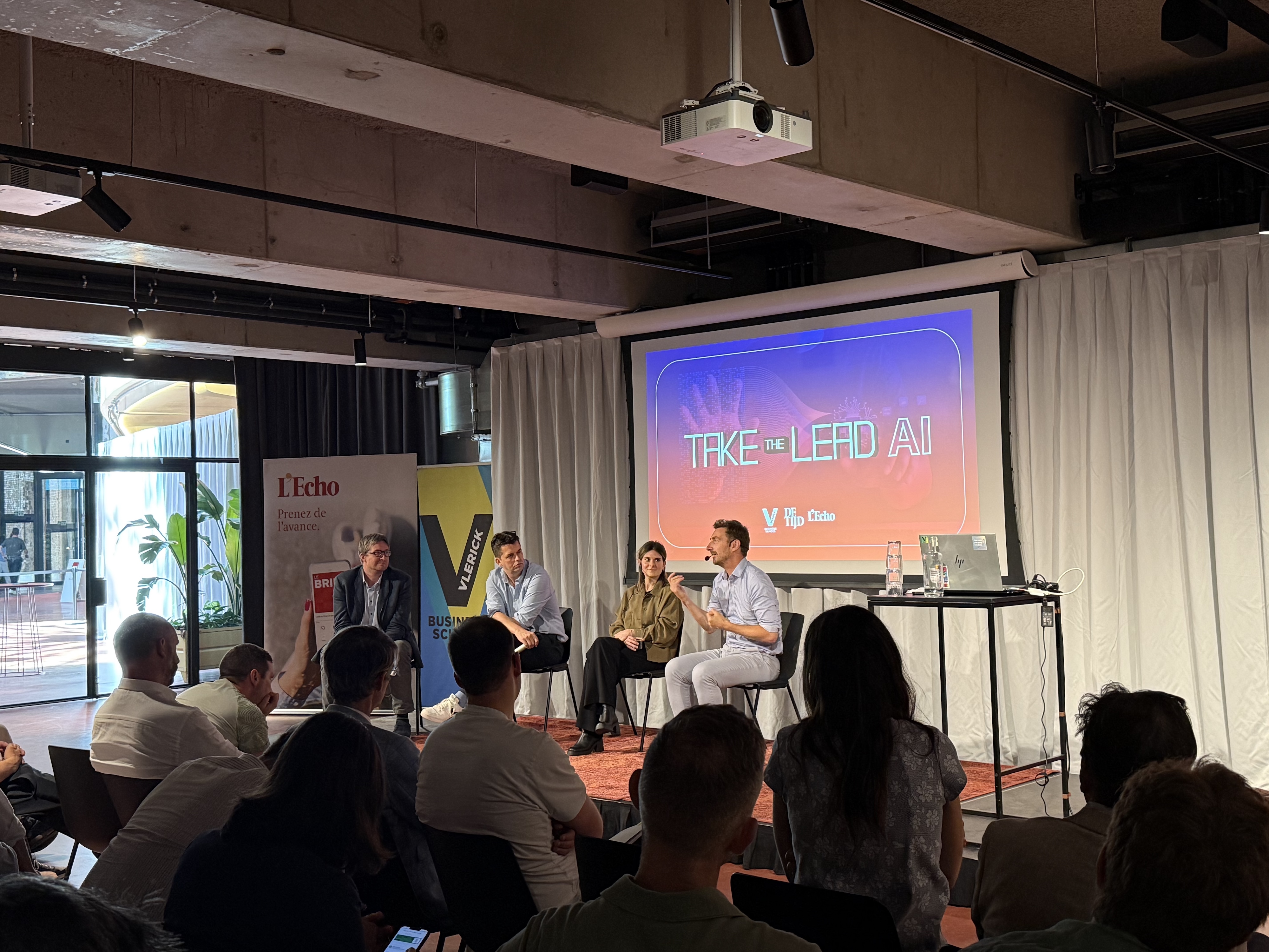 Four panellists on a low stage at a Take the Lead in AI event. The speaker on the right gestures into a headset mic, a woman in an olive jacket beside him. A large screen behind reads Take the Lead AI, with an audience watching nearby.