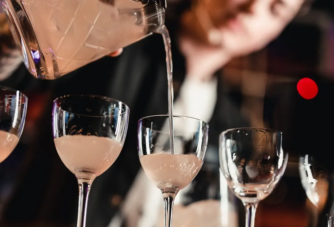 A bartender is pouring a whitish drink in a Nick & Nora glass. 