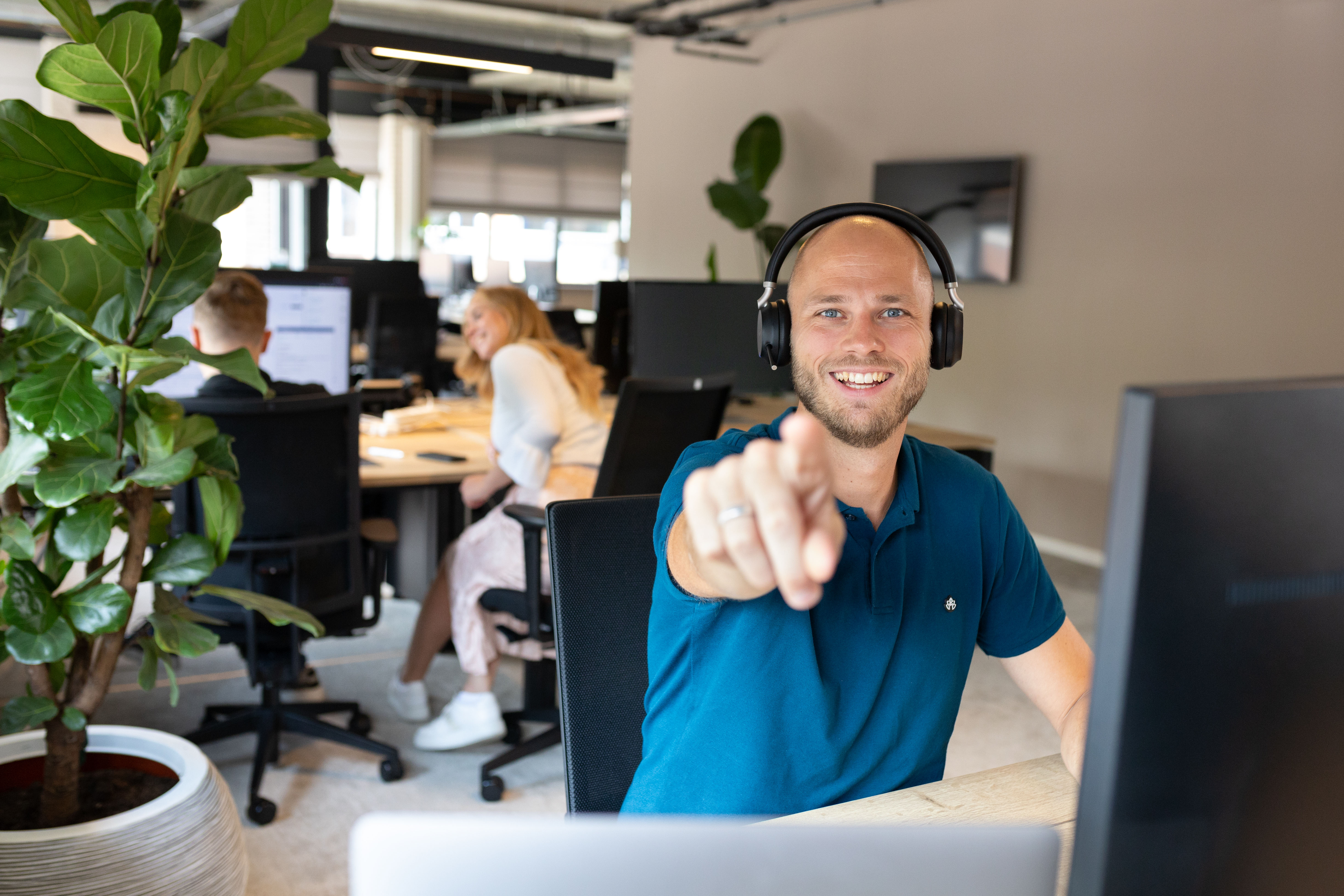 vPlan supportmedewerker met headset glimlacht en wijst naar de camera op het vPlan kantoor.