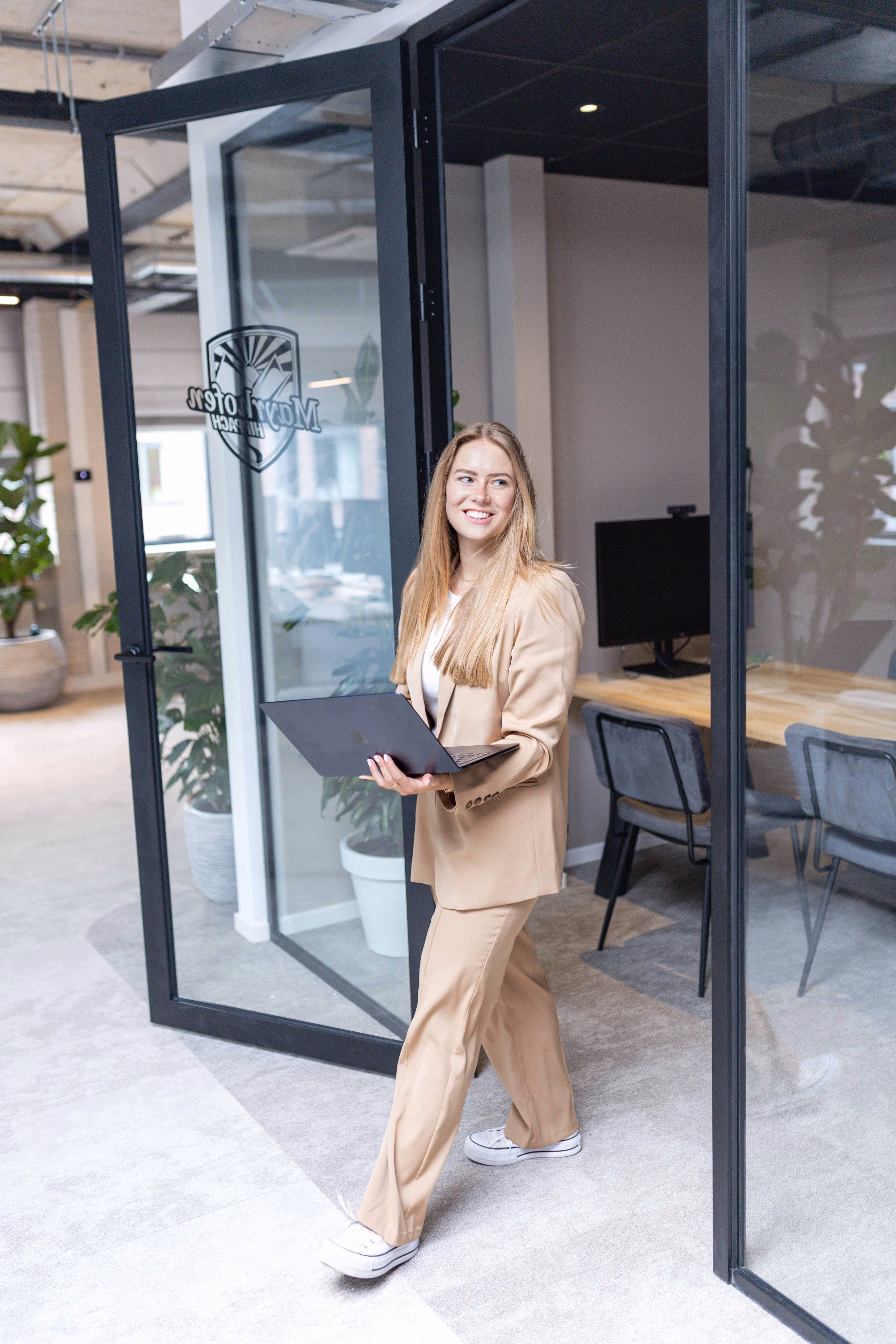 vPlan-medewerker in een beige pak loopt met een laptop en koffiebeker een vergaderruimte uit in het kantoor van vPlan.