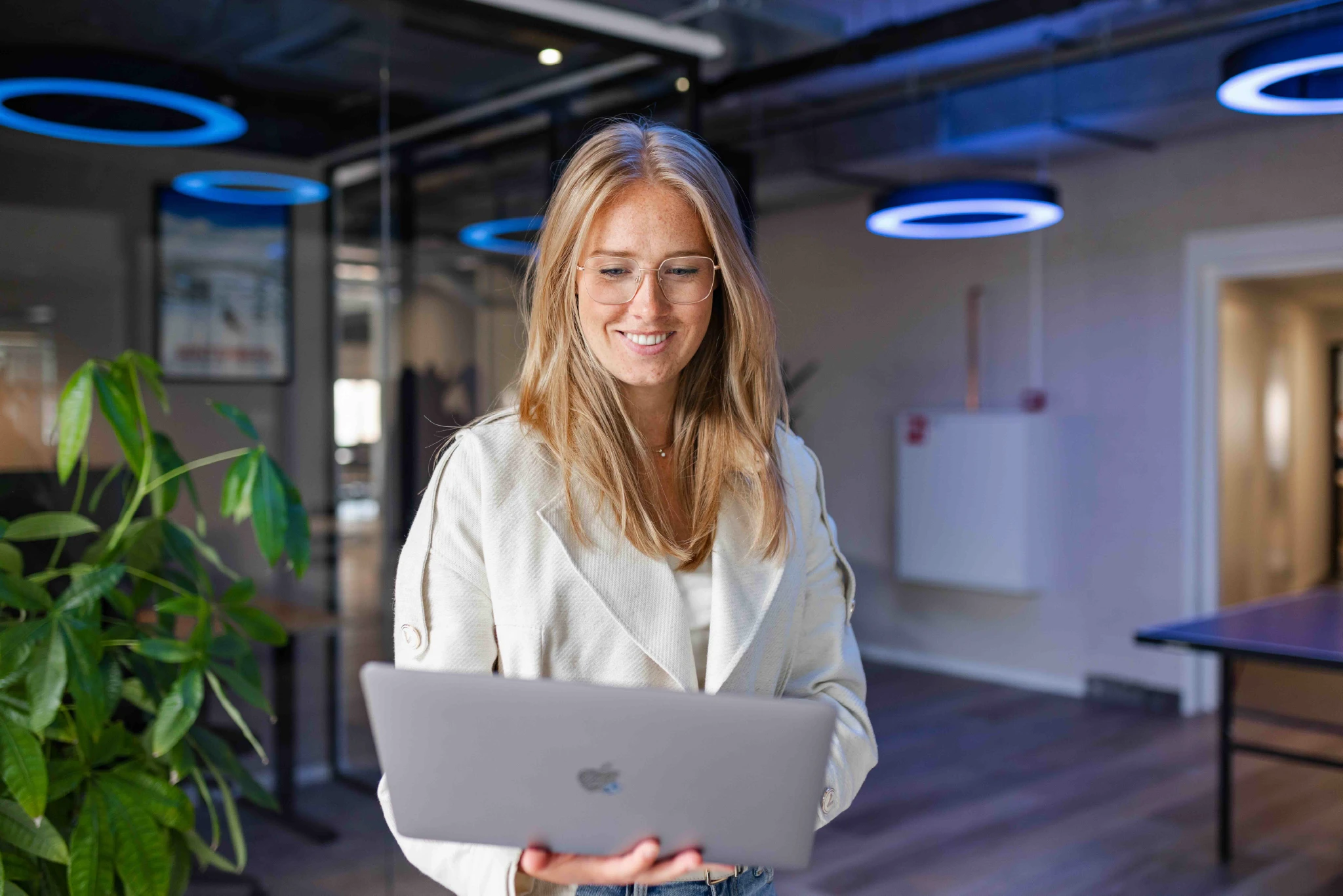 Vrouw in moderne kantooromgeving die glimlachend op haar laptop werkt, met focus op samenwerking en digitale productiviteit.