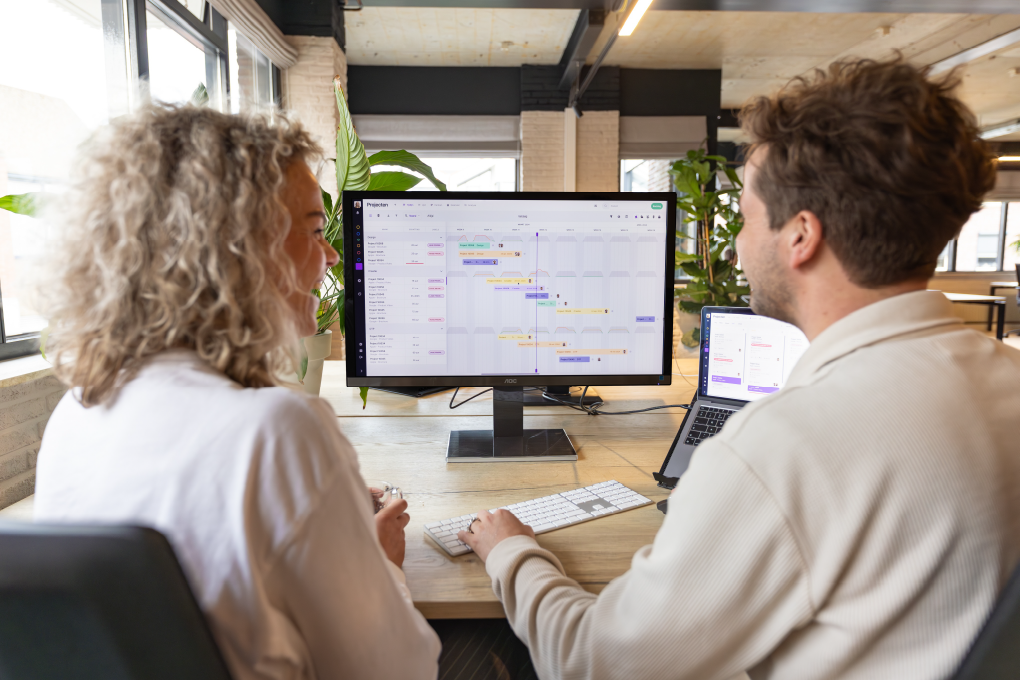 Two colleagues collaborating on a project plan displayed on a large monitor, showing the vPlan interface with tasks and timelines in a modern office setting.