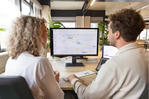 Two people looking at the vPlan planning software on a screen, showing tasks and a gantt chart overview.
