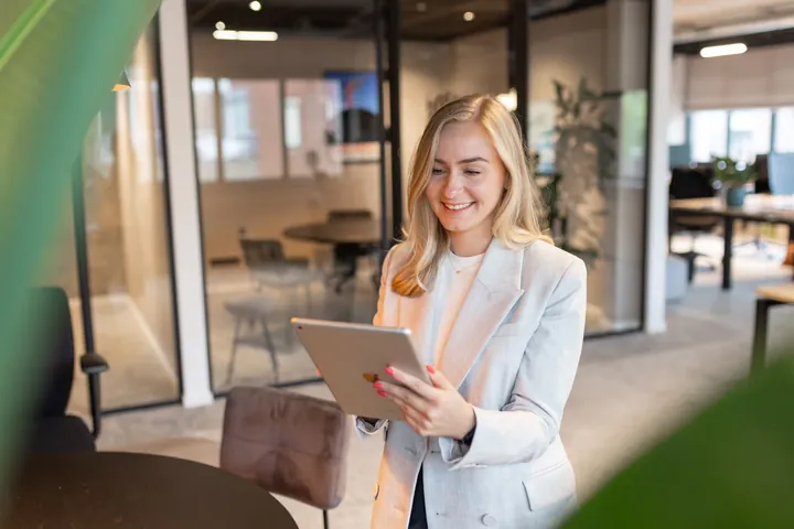 Smiling woman in light blazer using tablet in a modern office with glass walls and furniture.