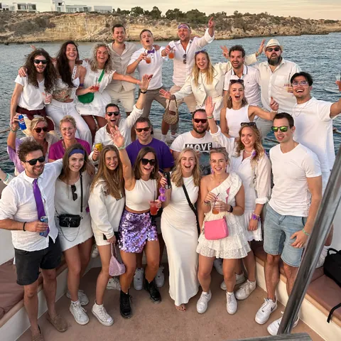 Group of adults smiling and posing on a boat with rocky coastline in the background at sunset.