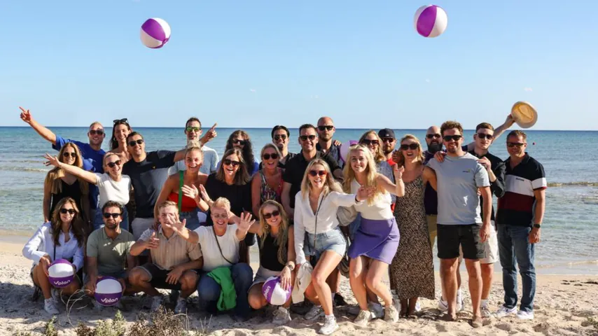 Group of people posing happily on a beach with purple and white beach balls and ocean in background.