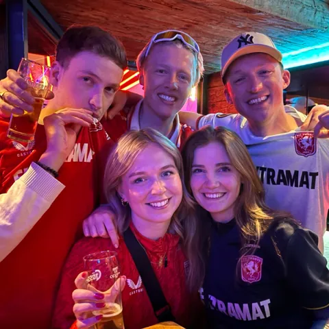 Group of five young adults smiling and holding drinks, wearing sports jerseys indoors with colorful lighting.