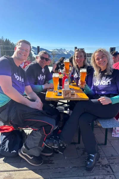 Four people in matching shirts sitting outdoors at a table with beers, snowy mountains in the background.