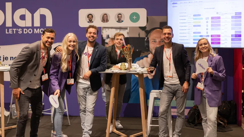 Six people in business casual at a booth with purple banners, smiling and posing for a group photo.
