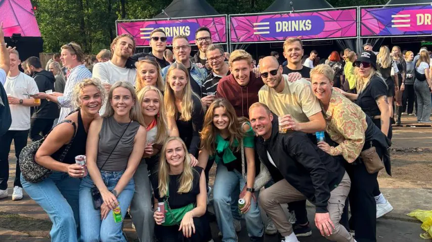 Group of friends smiling and posing for a photo at an outdoor festival near a drinks stand.