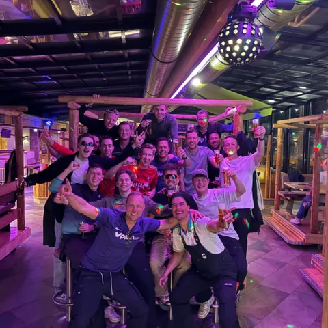 Group of men cheering and holding drinks inside a rustic bar with colorful lighting.