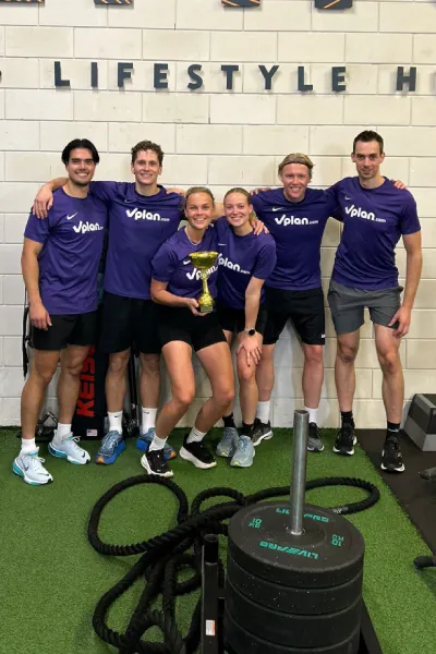 Five people in matching Vplan.com shirts posing with a trophy in a gym setting near weights and ropes.