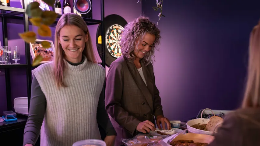 Two women smiling indoors while preparing food at a table with bread and spreads.
