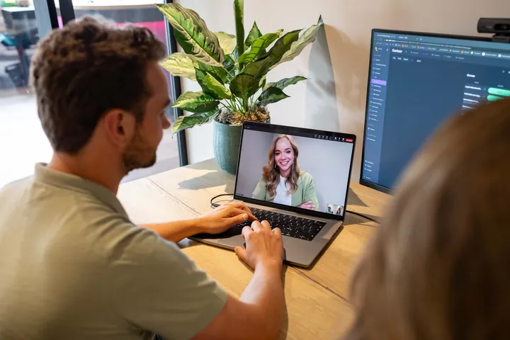 Man using laptop for video call with smiling woman wearing green jacket, sitting at wooden desk with plant.