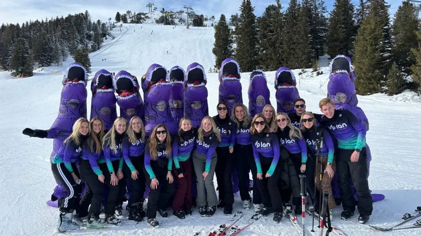 Group of people in purple dinosaur costumes and matching ski outfits posing on a snowy ski slope.