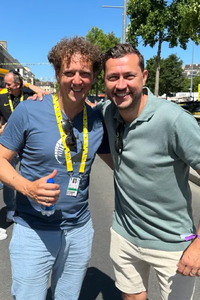 Two men smiling and posing outdoors on a sunny day, one giving a thumbs up and wearing a lanyard.