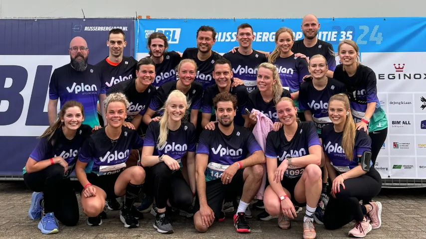 Group of 19 runners posing in front of event banners wearing matching Vplan shirts and race bibs.