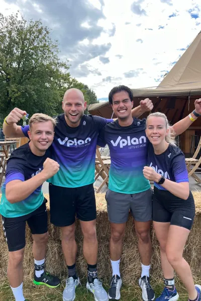 Four smiling runners in matching blue-green and black 'Vplan' shirts posing outdoors near hay bales.