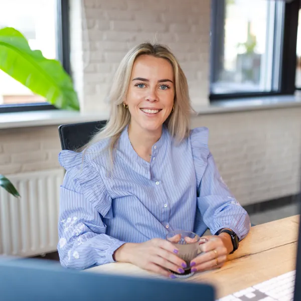 Smiling blonde woman in a blue blouse holding a cup, sitting at a wooden desk near windows.