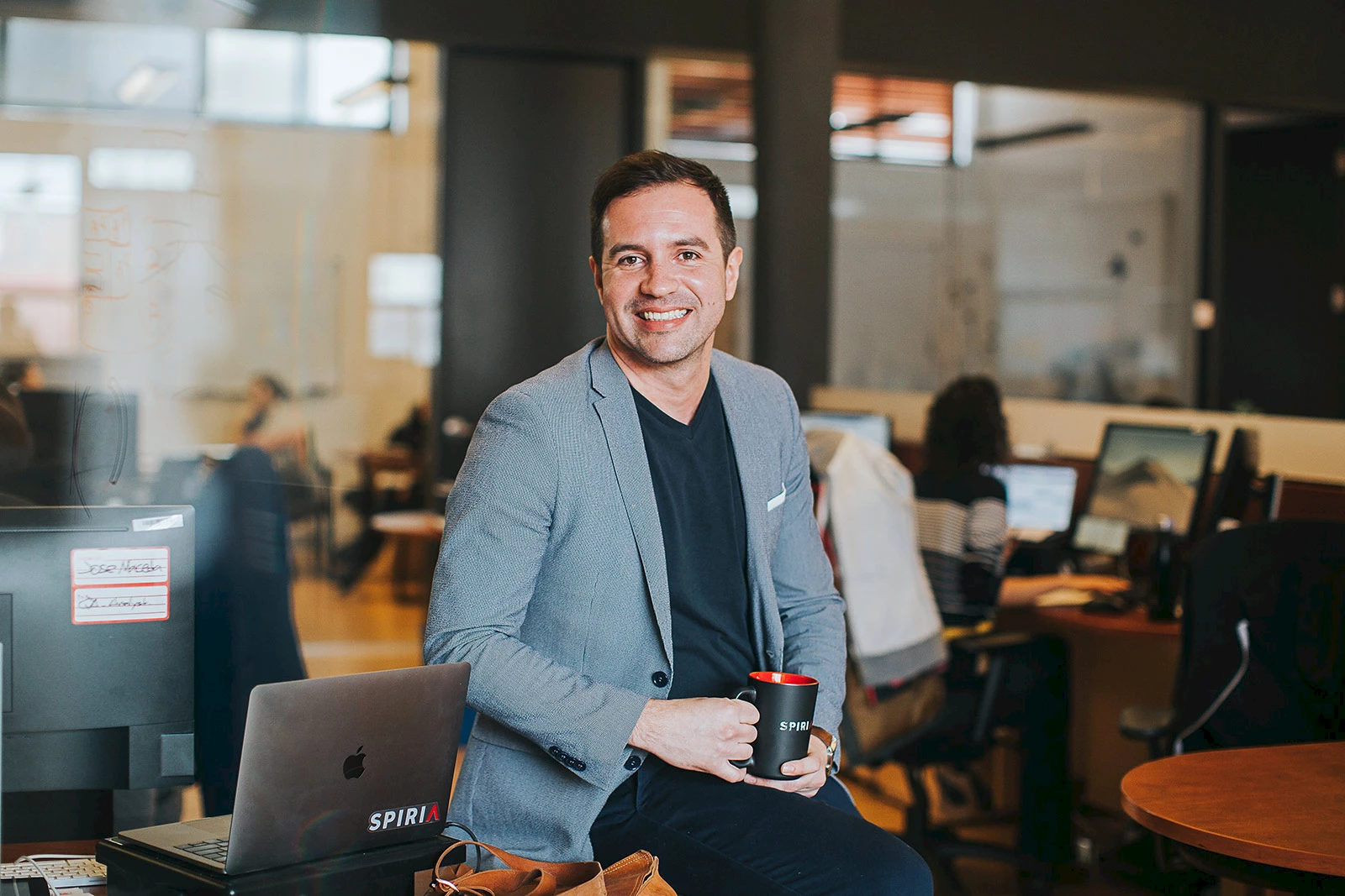 Smiling man in a gray blazer holding a black coffee mug sitting in a modern office with computers and colleagues working in the background.