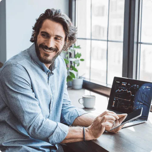 Smiling man using a tablet with a laptop in the background by a window.