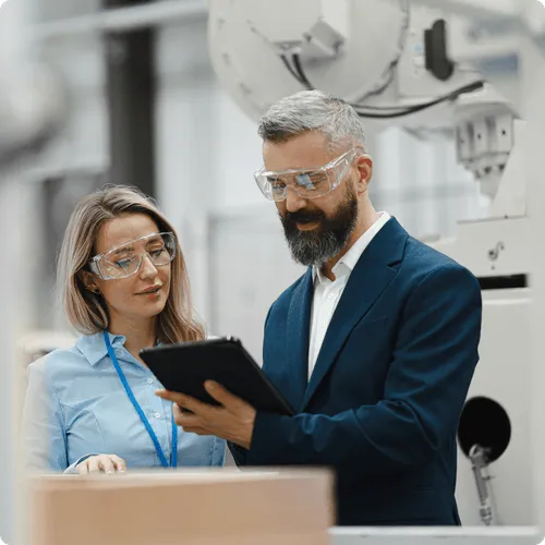 Two factory workers wearing safety glasses reviewing a tablet in an industrial setting.