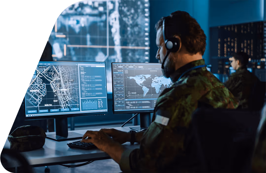 Military personnel in camouflage using dual monitors with maps and data in a command center.