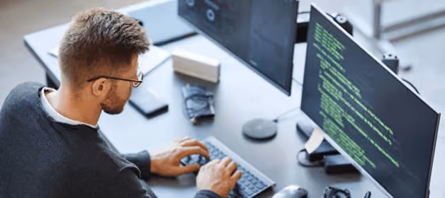 Man with glasses typing code on keyboard in front of dual monitors.