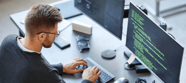 Man with glasses typing code on keyboard in front of dual monitors.