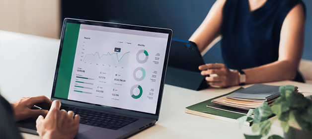 Une personne consulte des tableaux et des graphiques financiers sur son ordinateur portable tandis qu'une autre utilise une tablette à une table.