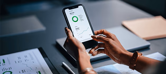 Une personne utilisant un smartphone sur lequel s'affiche une application financière comportant des tableaux et des graphiques, assise à un bureau où se trouvent des documents.