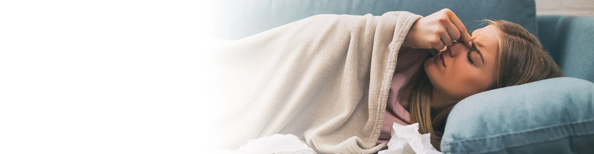 Woman lying on a couch wrapped in a blanket, pressing her fingers against her nose showing signs of sinus pain.