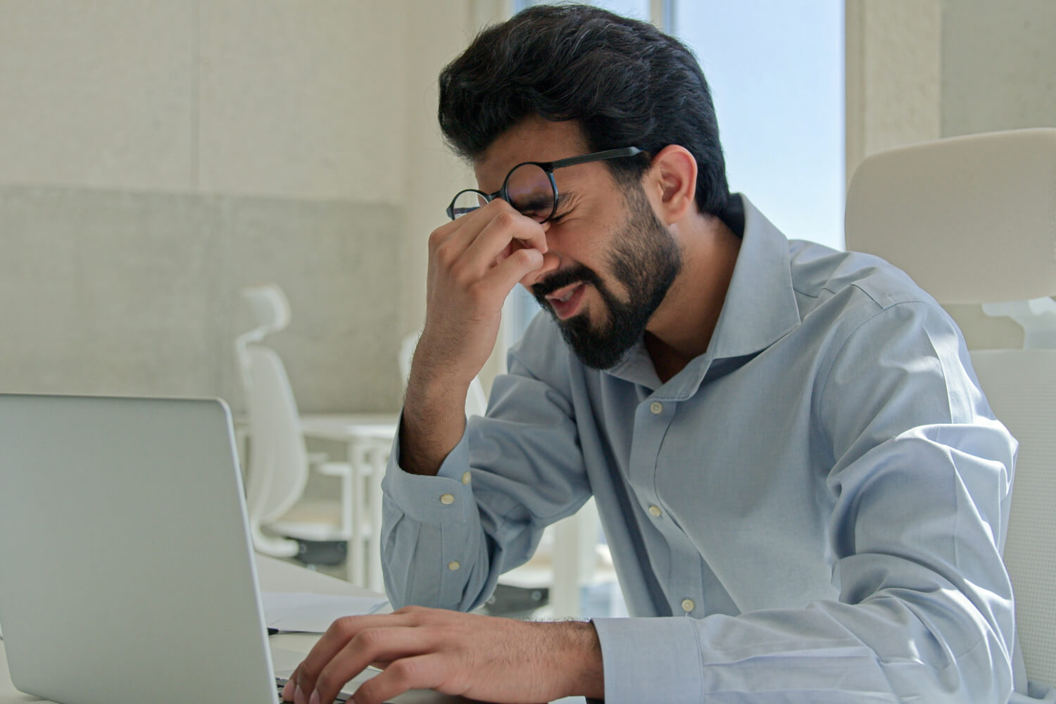 Man with glasses rubbing eyes while using laptop.