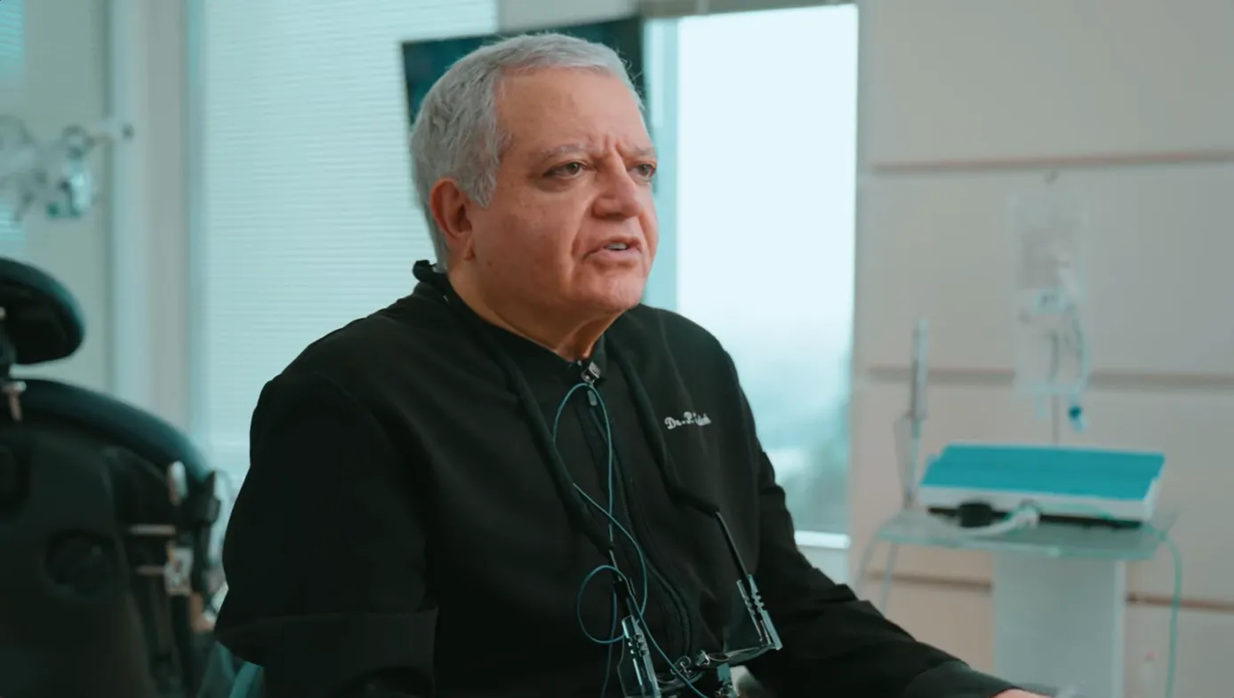 Middle-aged man with gray hair wearing a black medical coat seated in a clinical setting with medical equipment and a chair in the background.