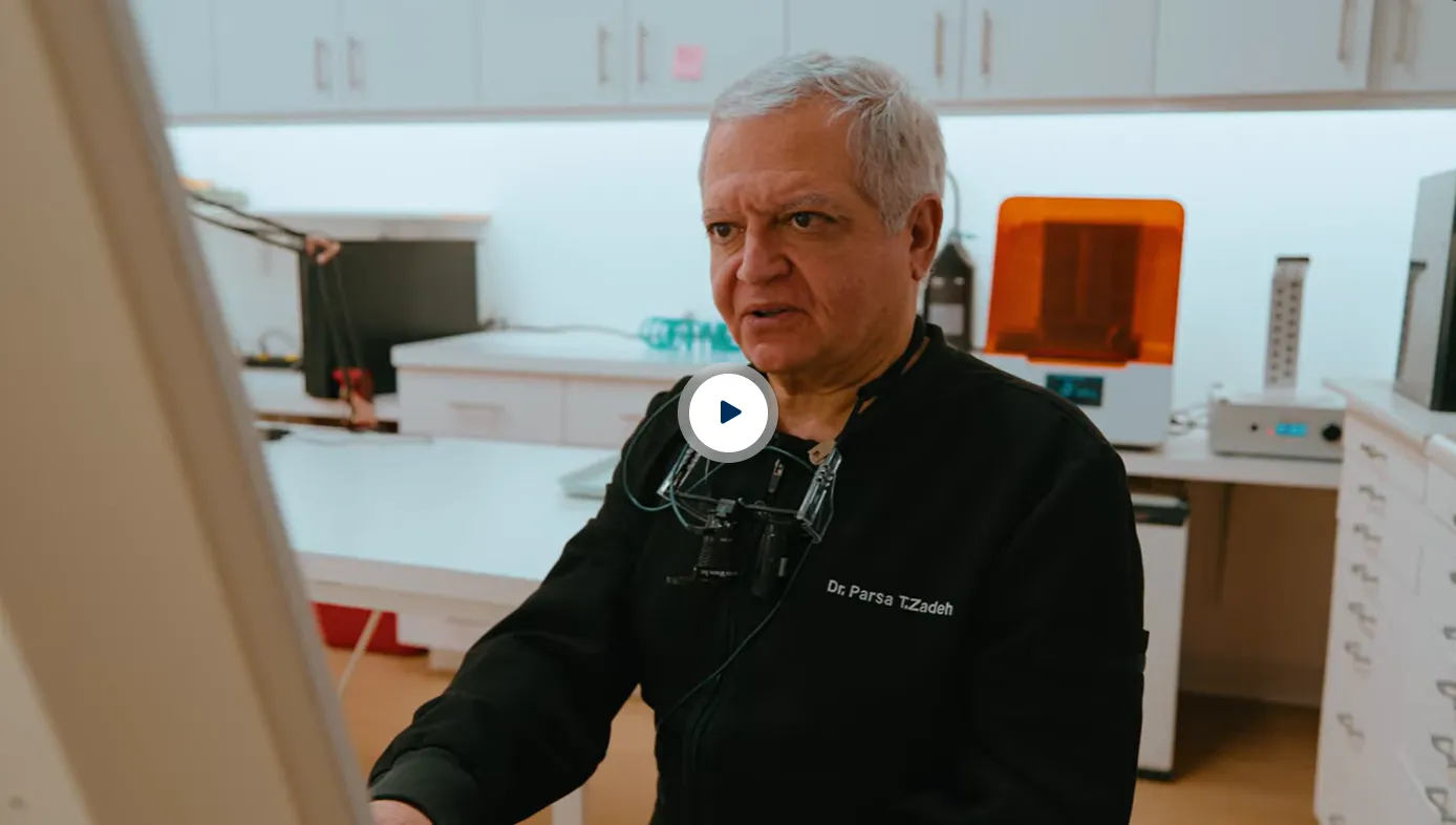 Older male dentist in black uniform with the name Dr. Parsa T.Zadeh working in a modern dental office with white cabinets and an orange 3D printer in the background.
