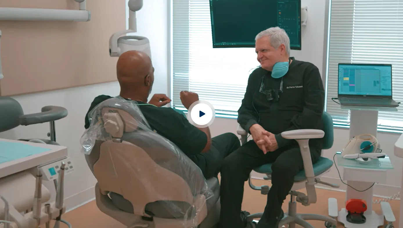 A dentist wearing black scrubs and a blue face mask sits on a swivel chair talking and smiling with a patient seated on a dental chair covered with protective plastic in a bright dental office with equipment and a laptop nearby.