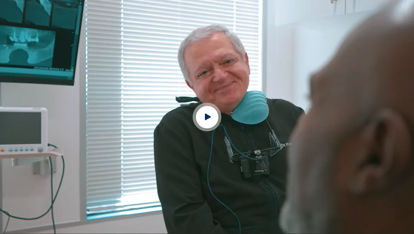 Smiling male dentist wearing a black jacket with dental magnifying glasses and a blue mask around his neck, sitting in a dental clinic with an X-ray displayed on a screen behind him facing a patient.