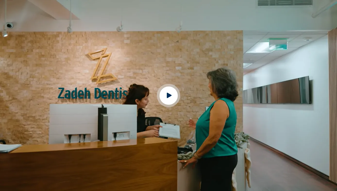 Reception area of Zadeh Dentistry with a receptionist handing a clipboard to a standing woman in a teal sleeveless top, against a background of a wooden wall and dental logo.